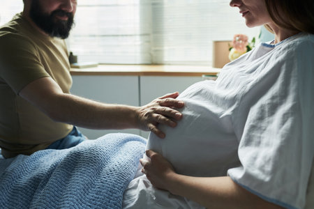 Caucasian pregnant woman sitting in hospital bed with middle aged Caucasian man gently touching her belly, both preparing for childbirth in medical setting, natural daylight entering roomの写真素材