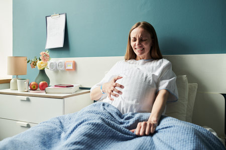 Caucasian young adult woman experiencing labor pain while lying in hospital bed, gripping blanket with tense expression, medical equipment and personal items visible on bedside tableの写真素材
