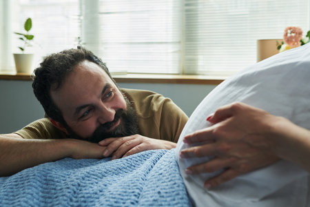 Middle aged Caucasian man smiling and resting head on hands while looking at pregnant woman touching belly, showing affectionate moment between expectant couple indoorsの写真素材
