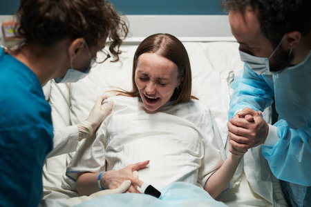 Caucasian young adult woman experiencing labor pain in hospital bed, supported by medical professional and Caucasian young adult man holding her hand during childbirth processの写真素材