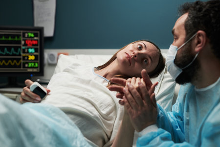 Caucasian young adult woman lying in hospital bed holding hands with Caucasian young adult man wearing medical mask and gown, medical monitor displaying vital signs in backgroundの写真素材