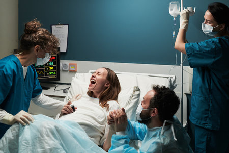 Caucasian young adult woman lying in hospital bed giving birth assisted by female medical staff while man holding her hand supporting during laborの写真素材