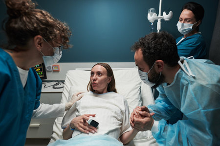 Caucasian young adult woman lying in hospital bed giving birth assisted by two female medical professionals while man holding her hand supporting herの写真素材