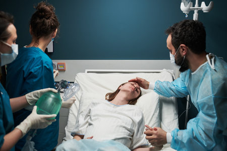 Caucasian young adult woman lying in hospital bed receiving medical attention from two female nurses and male doctor, healthcare professionals wearing protective masks and glovesの写真素材