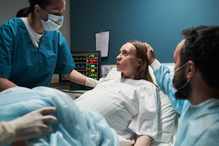 Caucasian young adult woman lying in hospital bed experiencing labor assisted by female nurse in scrubs while man supporting her during childbirth processの写真素材