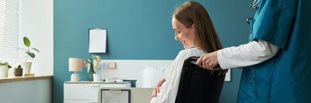 Caucasian young adult woman with disability sitting in wheelchair being assisted by female medical professional in clinic setting, both facing away from camera, smiling and interactingの写真素材