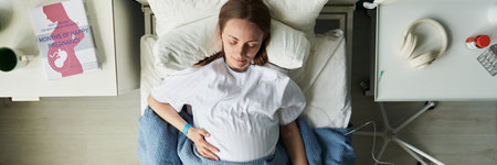 Caucasian young adult woman lying in hospital bed touching pregnant belly, relaxing with eyes closed, pregnancy book and headphones on bedside table, medical setting visibleの写真素材
