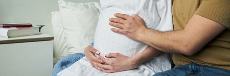 Caucasian pregnant woman sitting on bed being embraced by Caucasian man, both holding her belly with hands gently resting, showing intimate moment during pregnancy at homeの写真素材
