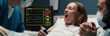 Caucasian young adult woman lying in hospital bed experiencing labor pain, gripping hand of supportive Caucasian man while medical professional assisting during childbirth processの写真素材