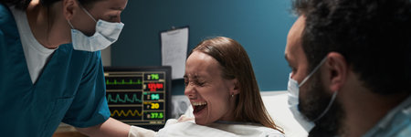 Caucasian young adult woman experiencing labor pain while lying in hospital bed, assisted by female nurse and bearded Caucasian young adult man, medical monitor displaying vital signsの写真素材