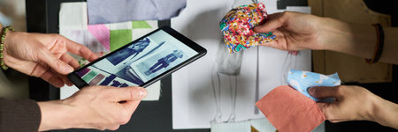 Caucasian woman holding tablet displaying fashion sketches while another Caucasian woman selecting colorful fabric swatches, collaborating on clothing design project at workspaceの写真素材
