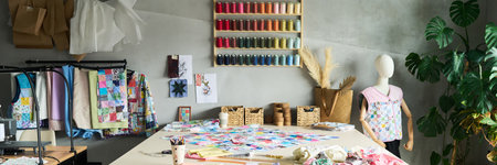 Sewing studio showing large worktable covered with colorful fabric pieces, mannequin displaying patchwork top, wall shelves holding spools of thread, sewing supplies organized in basketsの写真素材
