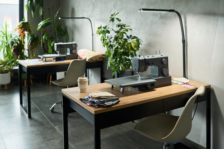 Empty sewing studio featuring two modern sewing machines on wooden desks with fabric pieces and coffee cup, indoor plants in background, contemporary workspace settingの写真素材