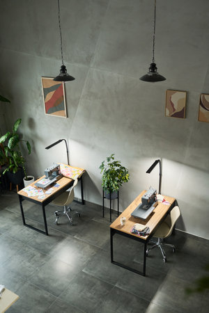 Empty modern workspace showing two sewing machines on wooden desks with fabric pieces, ergonomic chairs, potted plants, abstract wall art, overhead pendant lights, tiled floorの写真素材
