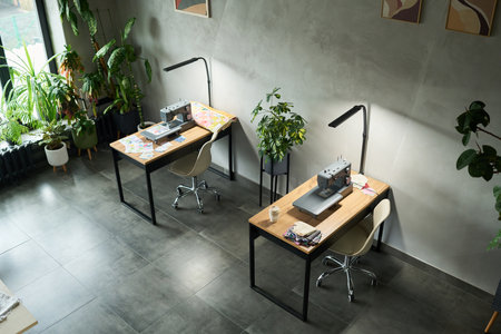 Two empty workstations with sewing machines positioned on wooden desks in modern studio, fabric and...