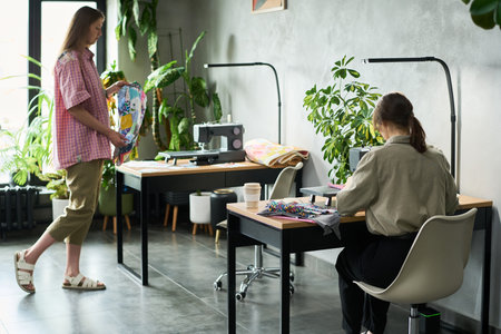 Caucasian young adult woman standing holding colorful fabric, while another Caucasian young adult woman sitting working at sewing machine in modern workspace with plants visibleの写真素材