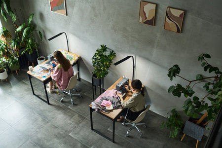 Two young adult Caucasian women sitting at separate desks working on creative projects, one drawing with colored pencils, other using sewing machine, surrounded by indoor plantsの写真素材