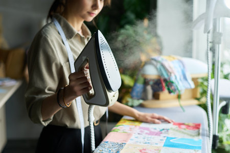 Young adult Caucasian woman ironing colorful fabric pieces on table, holding steam iron in hand, measuring tape around neck, standing in bright workspace with sewing materials visibleの写真素材