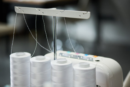Close up showing four large spools of thread mounted on industrial sewing machine, threads extending upward toward metal guide, focus on equipment used in textile productionの写真素材