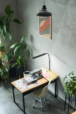 Modern workspace featuring sewing machine on wooden desk surrounded by green potted plants, colorful fabric pieces and paper patterns scattered across tabletop, empty chair positioned nearbyの写真素材