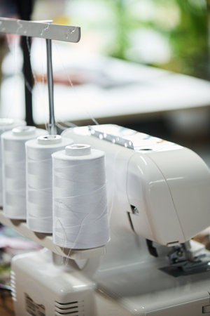Close up showing industrial overlock sewing machine with multiple large spools of thread positioned on table in workshop, focusing on equipment used for garment productionの写真素材