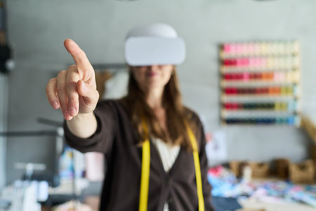 Caucasian young adult woman wearing virtual reality headset interacting with digital interface in creative workspace, extending hand forward and touching invisible screenの写真素材