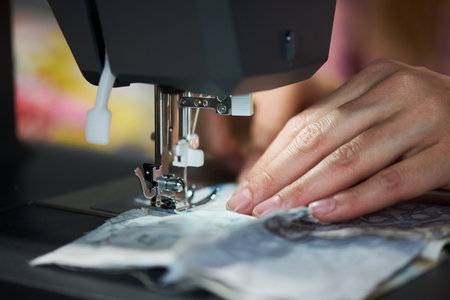 Caucasian young adult woman using sewing machine, hands guiding fabric under needle, focusing on precise stitching, close up of fingers and textile during garment creationの写真素材