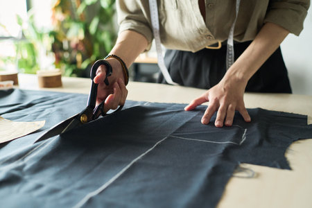 Caucasian young adult woman cutting fabric with large scissors on worktable, hands visible holding textile, measuring tape draped around neck, preparing garment patternの写真素材