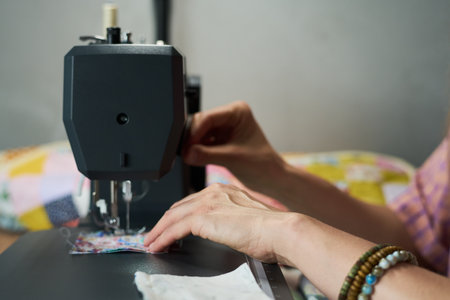 Caucasian young adult woman operating sewing machine, hands guiding fabric under needle, wearing beaded bracelets, focusing on precise stitching, colorful quilted textile in backgroundの写真素材