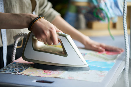 Caucasian young adult woman ironing colorful fabric pieces on ironing board, hand pressing steam iron while other hand smoothing material, close up of hands and armsの写真素材