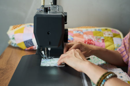 Caucasian woman sewing fabric with sewing machine, hands guiding material under needle, colorful patchwork quilt visible in background, focusing on precise stitchingの写真素材