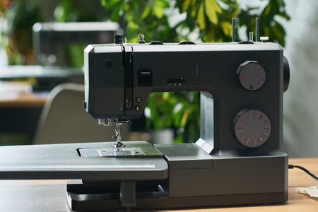 Modern sewing machine standing on table in workspace, metallic body featuring adjustment dials and extended platform, background showing blurred plants and furnitureの写真素材