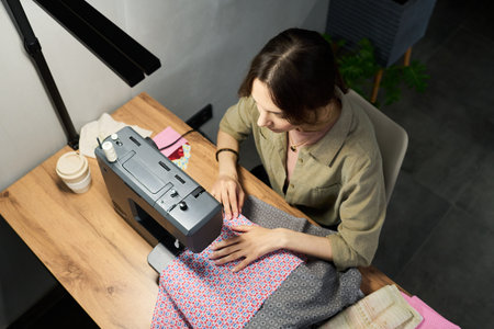 Young adult Caucasian woman using sewing machine, focusing on stitching patterned fabric at wooden desk, hands guiding textile, working in creative workspace, top view perspectiveの写真素材