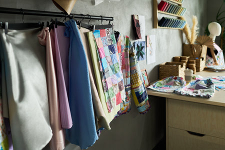 Colorful patchwork quilt and various fabrics hanging on rack in sewing studio, textile samples displayed on wall, sewing supplies and spools of thread arranged on wooden tableの写真素材