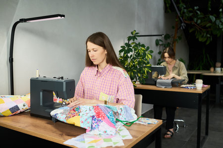 Caucasian young adult woman using sewing machine while working on colorful patchwork fabric at desk,...