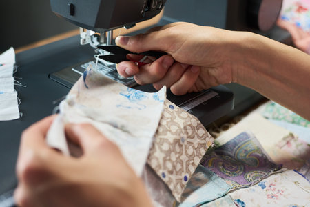 Caucasian young adult woman sewing fabric pieces with sewing machine, hands guiding patterned textiles under needle, focusing on precise stitching during textile crafting processの写真素材