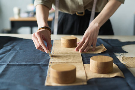 Young adult Caucasian woman marking fabric with tailor chalk while holding paper sewing pattern and...