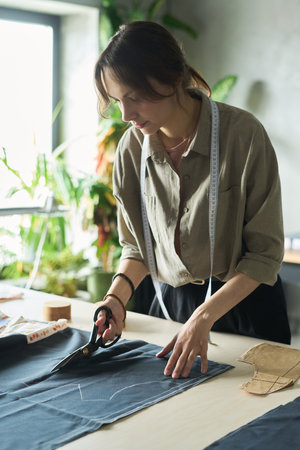 Caucasian young adult woman cutting fabric with scissors on worktable, measuring tape around neck, focusing on garment making process in bright studio with plants in backgroundの写真素材