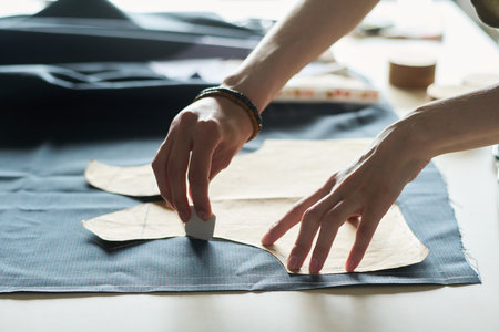 Caucasian young adult man marking fabric with tailor chalk while holding paper pattern, hands visible working on textile, preparing material for garment construction in workshopの写真素材