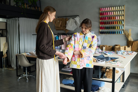 Caucasian young adult woman fitting colorful patchwork jacket on Caucasian girl in modern sewing studio, both standing near large worktable with sewing materials and thread spoolsの写真素材