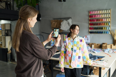 Caucasian young adult woman photographing Caucasian young adult woman standing near table in creative studio, colorful fabric samples hanging on wall, sewing materials visible in backgroundの写真素材