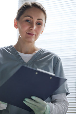 Vertical medium portrait of Caucasian woman working as doctor in hospital holding clipboard with medical record checking health state of patientの写真素材