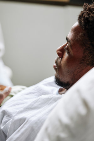 Vertical medium close-up of young Black man lying in hospital bed in ward thinking about somethingの写真素材