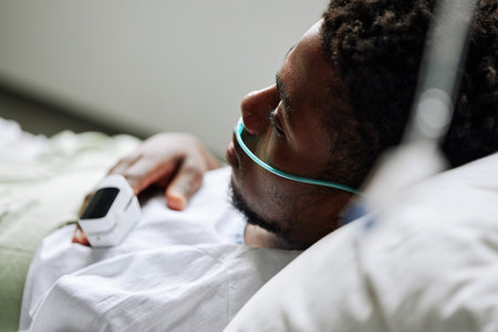 Medium close-up of young Black man with nasal cannula and pulse oximeter on finger lying in hospital bed in ward thinking of somethingの写真素材