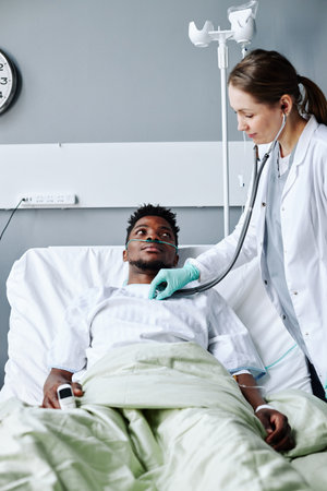 Caucasian woman wearing lab coat working in modern hospital checking health state of young Black man using stethoscopeの写真素材