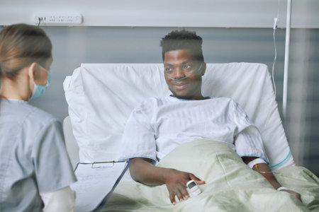 Unrecognizable Caucasian doctor wearing mask on face asking questions to young Black patient to check his health state in hospital wardの写真素材