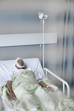 Through shutters shot of young African American man spending days in hospital ward after head injury sleeping while getting IV therapyの写真素材