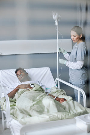 Through shutters shot of young African American man with nasal cannula lying in hospital bed looking away, Caucasian nurse setting IV line for himの写真素材