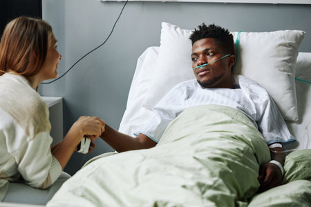 Horizontal shot of Caucasian woman visiting her African American friend in hospital ward holding his hand trying to cheer him upの写真素材