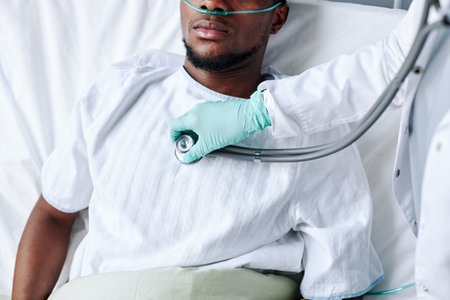 High angle view of unrecognizable doctor using stethoscope to listen to heart and lungs sounds of young Black man lying in hospital bedの写真素材
